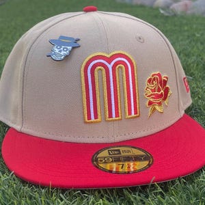May include: A tan baseball cap with a red brim and a red button on top. The cap features a large red and gold letter "M" patch, a red rose patch, and a skull pin. The cap has a "New Era" sticker on the brim.