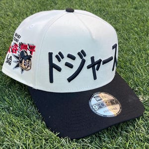 Puede incluir: Gorra de béisbol blanca y negra con la palabra "Dodgers" en caracteres japoneses bordada en la parte delantera. La gorra presenta un gráfico de dibujos animados y texto en el lateral, y una pegatina ajustable "9FORTY".