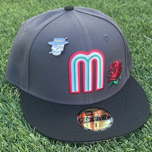 May include: A gray baseball cap with a black brim, featuring a pin of a skull wearing a hat and a colorful "M" patch. A red rose and a "59FIFTY" sticker are also visible. The hat size is 7 1/8.