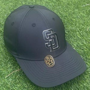May include: Black baseball cap with a silver "SD" logo and a gold pin featuring a skull design. The cap is on a green grassy surface.