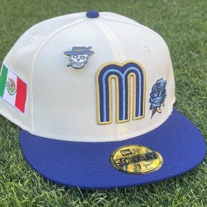 May include: Cream-colored baseball cap with a blue brim and top button. Features a Mexican flag patch, a skull pin, and a blue rose patch. The letter "M" is embroidered in blue and gold. The hat size is 7 1/2.