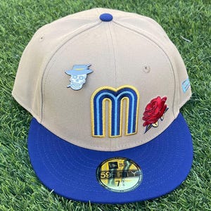 May include: A tan baseball cap with a blue brim. The cap features a skull pin with a hat, a rose, and a stylized "M" logo in blue and gold. The cap has a "59FIFTY" sticker and is size 7 3/8.