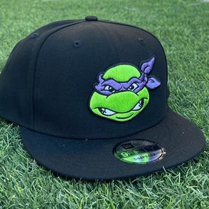 Puede incluir: Gorra de béisbol negra con cierre a presión, con una imagen bordada en verde y morado de un personaje de las Tortugas Ninja. La gorra tiene una visera plana y una pegatina New Era 9FIFTY Snapback.