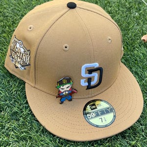 May include: A tan baseball cap with the San Diego Padres logo in white and black. The cap has a black button on top and a 7 3/8 size sticker. A pin of a cartoon character is attached to the front.