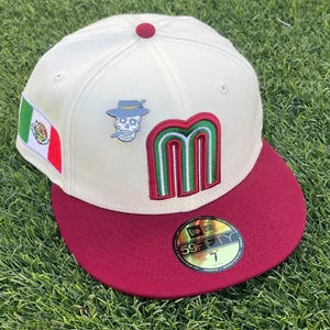 May include: Cream-colored baseball cap with a maroon brim and a red button on top. Features a Mexican flag patch, a skull pin with a hat, and a green and red embroidered "M" logo. The hat is a 59FIFTY fitted style.