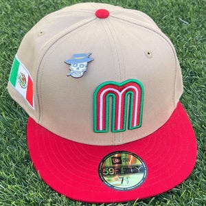 May include: Tan and red baseball cap with a flat brim. The cap features a Mexican flag patch, a skull pin wearing a hat, and a large letter "M" in green, red, and white. The cap has a red button on top and a "59FIFTY" sticker.