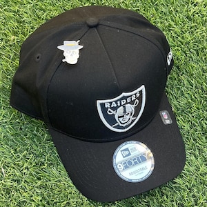 Op de afbeelding: Zwarte verstelbare baseballcap met het Raiders-logo in wit en zilver. Een schedelspeld met een hoed en een sigaret is bevestigd. De cap heeft een gebogen klep en een 9FORTY-sticker.