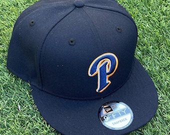 Pode incluir: Boné de beisebol azul marinho com aba curva. O boné apresenta uma letra "P" estilizada em azul com contorno laranja. Um adesivo "9FIFTY SNAPBACK" está na aba. O boné está sobre um fundo de grama verde.