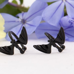 Pu&ograve; includere: Due orecchini a forma di pipistrello neri. Gli orecchini sono in metallo e hanno un design a spirale. Sono mostrati su uno sfondo bianco con petali di fiori gialli e blu.