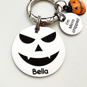May include: A silver dog tag featuring a black and white jack-o'-lantern face with the name "Bella" engraved on it. The tag is attached to a silver keyring with a small orange pumpkin charm and a silver tag that reads "I'm Microchipped".