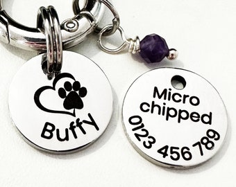 May include: Silver dog tag with a heart and paw print design, the name "Buffy" engraved on it. A second silver tag reads "Micro chipped" and includes a phone number. A purple bead is attached to the tags.
