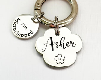 May include: Silver dog tag keychain with a paw print design. The tag is engraved with the name "Asher" and a small flower. A smaller silver tag is attached to the keychain and reads "I'm microchipped".
