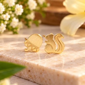 May include: A pair of gold-toned stud earrings featuring a brushed metal finish. One earring is shaped like an acorn with a geometric triangular pattern on its cap, and the other earring is shaped like a squirrel with a curved, textured tail.
