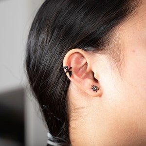 Pu&ograve; includere: Una persona con i capelli neri che indossa un braccialetto per orecchie a forma di pipistrello nero e un orecchino a forma di ragno nero.