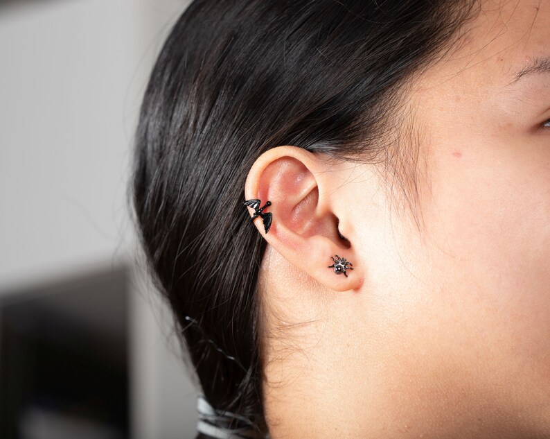 Pu&ograve; includere: Una persona con i capelli neri che indossa un braccialetto per orecchie a forma di pipistrello nero e un orecchino a forma di ragno nero.