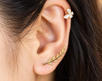 May include: A gold-toned ear crawler earring shaped like a leafy branch with textured details, paired with a small gold-toned butterfly stud earring featuring embedded clear stones. Both pieces are worn on the ear, showcasing a metallic finish.
