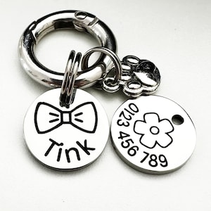 May include: Two silver dog tags with black engravings. One tag features a bow tie and the name "Tink". The other tag has a flower and the phone number "0123 456 789".