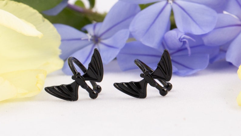 Pu&ograve; includere: Due orecchini a forma di pipistrello neri. Gli orecchini sono in metallo e hanno un design a spirale. Sono mostrati su uno sfondo bianco con petali di fiori gialli e blu.