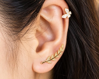 May include: A gold-toned ear crawler earring shaped like a leafy branch with textured details, paired with a small gold-toned butterfly stud earring featuring embedded clear stones. Both pieces are worn on the ear, showcasing a metallic finish.
