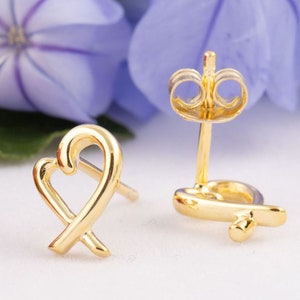 May include: A pair of gold stud earrings. Each earring has a heart-shaped design with an intertwined X. The earrings are displayed on a white surface with blurred purple flowers in the background.