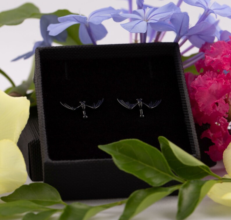 Pu&ograve; includere: Un paio di orecchini a forma di pipistrello neri in una scatola di gioielli in velluto nero. Gli orecchini sono piccoli e delicati, con un design di pipistrello dettagliato.