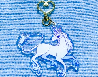 Puede incluir: Un llavero de unicornio blanco con un contorno azul y un cierre dorado en forma de corazón. El unicornio está mirando hacia la derecha y tiene una melena y una cola fluidas.
