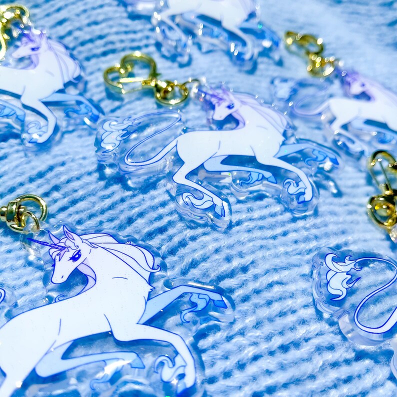 May include: A set of five clear acrylic keychains featuring a white unicorn design. The unicorns are in various poses and have a blue iridescent shimmer.