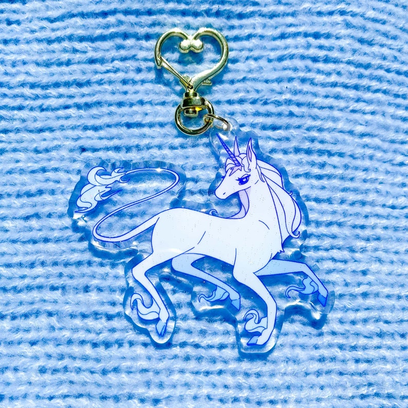 May include: A white unicorn keychain with a blue outline and a gold heart-shaped clasp. The unicorn is facing right and has a flowing mane and tail.