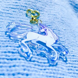 May include: A white unicorn keychain with a gold key ring. The unicorn is depicted in a running pose with a flowing mane and tail. The keychain is made of clear acrylic with a glittery finish.