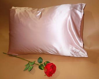 May include: A rectangular pillowcase in a soft, pale pink color with a smooth, lustrous finish. The fabric features a subtle embroidered logo in the bottom right corner. A single artificial red rose with green leaves lies in front of the pillowcase.
