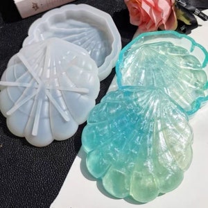 May include: Two clear resin seashell-shaped boxes. One box is a light blue color and the other is a translucent green color. Both boxes have a raised seashell design.