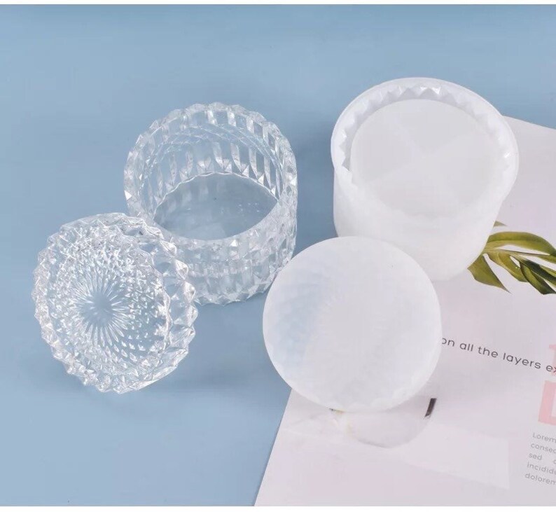May include: Clear, faceted glass jar with lid, next to a white, round, frosted object and a white container. The jar is open, revealing its interior. The objects are on a light blue surface.