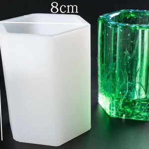 May include: A white, hexagonal-shaped silicone mold, 11 cm tall and 8 cm wide, next to a green, hexagonal-shaped resin casting. The resin casting has a textured surface and is filled with bubbles.