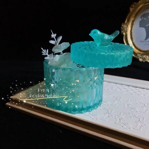 May include: A turquoise resin jewelry box with a faceted design. The lid is topped with a bird figurine. The box is open, revealing white decorative elements inside. The text "FYFM Resin&Molds" is visible on the base.