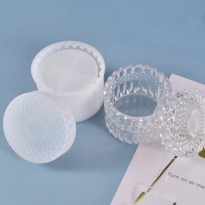 May include: Two decorative containers. One is a clear, faceted glass jar with a lid. The other is a white, round container with a lid. Both lids have a textured, geometric pattern. The background is a light blue.