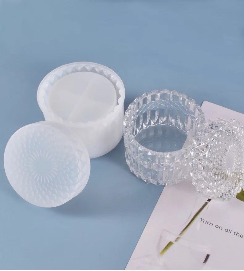 May include: Two decorative containers. One is a clear, faceted glass jar with a lid. The other is a white, round container with a lid. Both lids have a textured, geometric pattern. The background is a light blue.