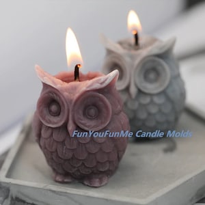 May include: Two owl-shaped candles, one in a light purple color and the other in a gray color. Both candles are lit and have a flame.