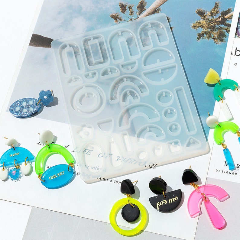 May include: A clear silicone mold with multiple shapes for making earrings. The mold is on a white surface with colorful resin earrings nearby. The earrings are in various shapes, including arches, circles, and half-circles. The earrings are in shades of blue, green, and pink.