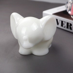 Puede incluir: Una figurita de elefante de cer&aacute;mica blanca con un acabado liso y brillante. El elefante tiene orejas grandes y una trompa enroscada.