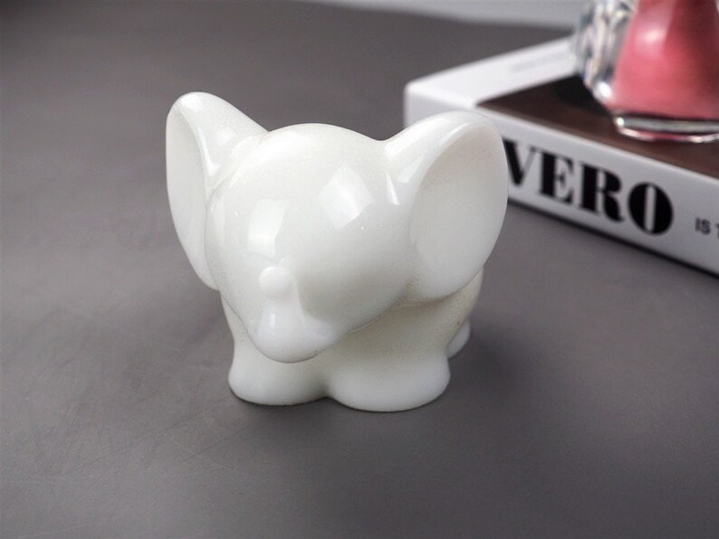 Puede incluir: Una figurita de elefante de cer&aacute;mica blanca con un acabado liso y brillante. El elefante tiene orejas grandes y una trompa enroscada.