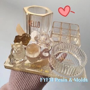 May include: A collection of clear resin molds, including a hexagonal pen holder with "HELLO" text, a multi-compartment organizer, and a ribbed ring dish. The set includes decorative resin elements like a flower, a heart, and a pig figurine, all on a gold glitter base.