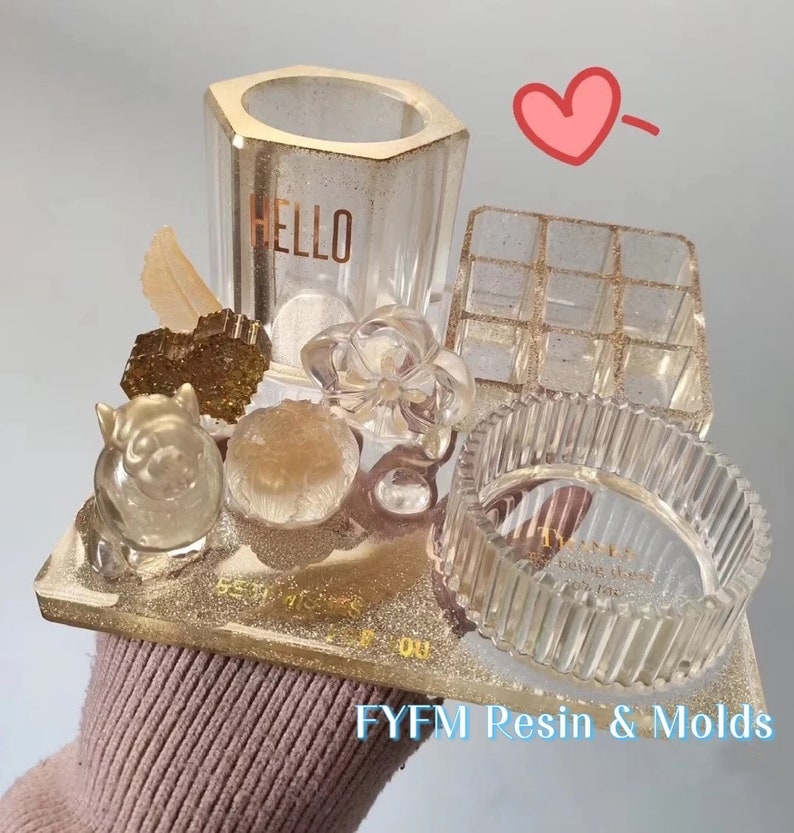 May include: A collection of clear resin molds, including a hexagonal pen holder with "HELLO" text, a multi-compartment organizer, and a ribbed ring dish. The set includes decorative resin elements like a flower, a heart, and a pig figurine, all on a gold glitter base.