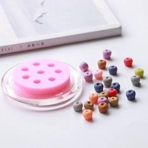 Op de afbeelding: Een roze siliconen mal met negen ronde holtes voor het maken van kleine appelvormige kralen. Een verscheidenheid aan kleurrijke polymer clay kralen zijn verspreid rond de mal.