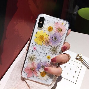 May include: A clear phone case with a glittery background and pressed flowers. The flowers are yellow, pink, purple, and white.