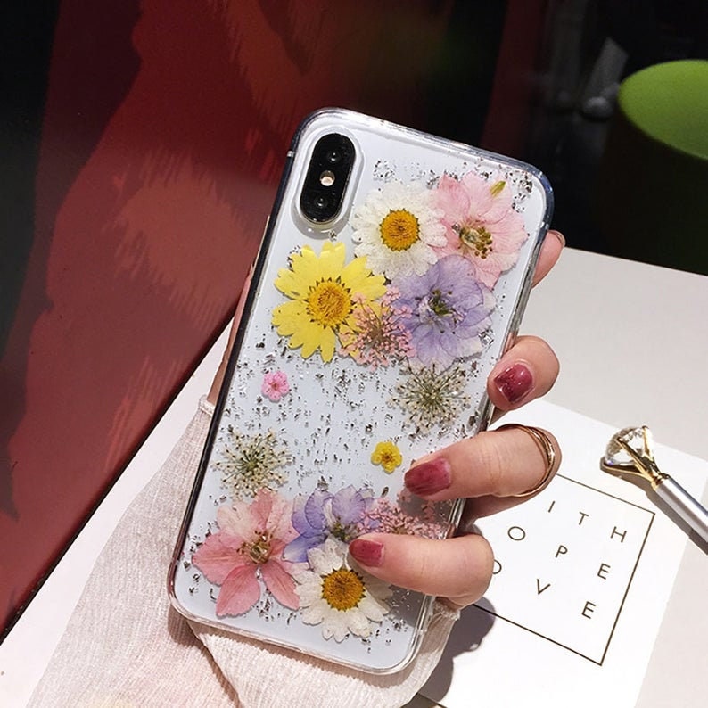 May include: A clear phone case with a glittery background and pressed flowers. The flowers are yellow, pink, purple, and white.