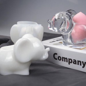 Puede incluir: Dos recipientes de cer&aacute;mica blanca con forma de elefante, uno con un recipiente de vidrio transparente con forma de elefante que contiene esponjas de maquillaje rosas. El recipiente de vidrio est&aacute; colocado sobre un libro con la palabra "Company" impresa en la portada.
