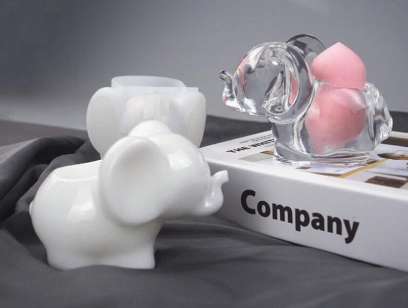 Puede incluir: Dos recipientes de cer&aacute;mica blanca con forma de elefante, uno con un recipiente de vidrio transparente con forma de elefante que contiene esponjas de maquillaje rosas. El recipiente de vidrio est&aacute; colocado sobre un libro con la palabra "Company" impresa en la portada.