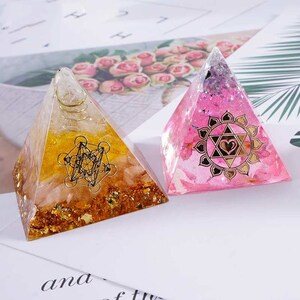 Pu&ograve; includere: Due piramidi di cristallo con accenti dorati. Una piramide &egrave; rosa con un simbolo di cuore dorato e l'altra &egrave; gialla con un design geometrico dorato.