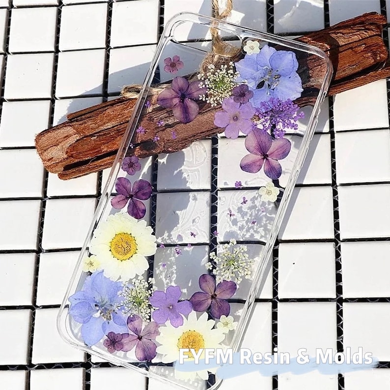 May include: A clear phone case with pressed flowers inside. The flowers are purple, blue, and white. The case is on a white tiled surface. The text "FYFM Resin & Molds" is visible in the bottom right corner.
