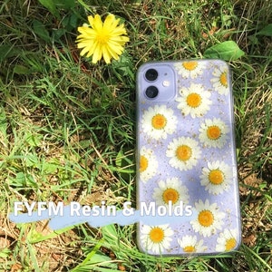 May include: A clear phone case with a purple background and a pattern of white daisies and gold glitter. The case is lying on a bed of green grass.